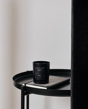 A black candle with a label sits on top of a closed book, both placed on a round black table. The table is next to a tall, dark curtain and a plain, light-colored wall.