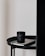 A black candle with a label sits on top of a closed book, both placed on a round black table. The table is next to a tall, dark curtain and a plain, light-colored wall.