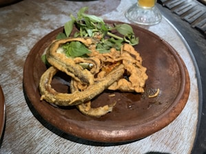 A brown ceramic plate holds several pieces of deep-fried food alongside fresh green herbs. The food appears crispy and golden, curling and overlapping on the plate. A glass with a beverage is partially visible in the background.