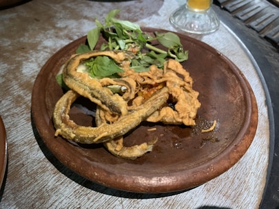 A brown ceramic plate holds several pieces of deep-fried food alongside fresh green herbs. The food appears crispy and golden, curling and overlapping on the plate. A glass with a beverage is partially visible in the background.