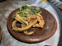 A brown ceramic plate holds several pieces of deep-fried food alongside fresh green herbs. The food appears crispy and golden, curling and overlapping on the plate. A glass with a beverage is partially visible in the background.