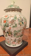 A large, ornate porcelain vase with intricate hand-painted scenes and patterns covering its surface. The artwork includes depictions of landscapes, flora, fauna, and possibly mythical creatures, set within panels. The colors are vibrant, featuring greens, reds, and whites. The lid has additional decorative elements, including a sculpted creature on top.