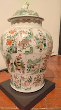 A large, ornate porcelain vase with intricate hand-painted scenes and patterns covering its surface. The artwork includes depictions of landscapes, flora, fauna, and possibly mythical creatures, set within panels. The colors are vibrant, featuring greens, reds, and whites. The lid has additional decorative elements, including a sculpted creature on top.