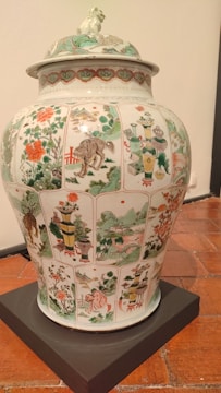 A large, ornate porcelain vase with intricate hand-painted scenes and patterns covering its surface. The artwork includes depictions of landscapes, flora, fauna, and possibly mythical creatures, set within panels. The colors are vibrant, featuring greens, reds, and whites. The lid has additional decorative elements, including a sculpted creature on top.