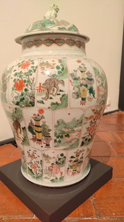 A large, ornate porcelain vase with intricate hand-painted scenes and patterns covering its surface. The artwork includes depictions of landscapes, flora, fauna, and possibly mythical creatures, set within panels. The colors are vibrant, featuring greens, reds, and whites. The lid has additional decorative elements, including a sculpted creature on top.