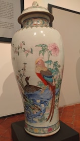 A large, decorative porcelain vase with intricate hand-painted designs featuring colorful birds, elaborate floral patterns, and a detailed landscape scene. The vase has a lid and is displayed on a black pedestal in a gallery setting.