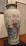 A large, decorative porcelain vase with intricate hand-painted designs featuring colorful birds, elaborate floral patterns, and a detailed landscape scene. The vase has a lid and is displayed on a black pedestal in a gallery setting.