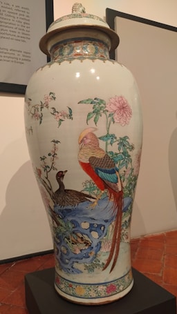A large, decorative porcelain vase with intricate hand-painted designs featuring colorful birds, elaborate floral patterns, and a detailed landscape scene. The vase has a lid and is displayed on a black pedestal in a gallery setting.
