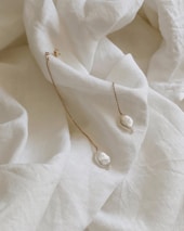 A delicate pair of gold chain earrings with baroque pearls is laid upon a soft, crumpled white fabric. The fabric has a textured appearance, creating a gentle, luxurious background for the earrings. The earrings feature small, irregular pearls at the end of fine, golden chains that reflect light subtly.