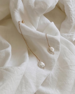 A delicate pair of gold chain earrings with baroque pearls is laid upon a soft, crumpled white fabric. The fabric has a textured appearance, creating a gentle, luxurious background for the earrings. The earrings feature small, irregular pearls at the end of fine, golden chains that reflect light subtly.