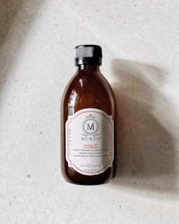 A plastic bottle with a dark amber color and a black cap is placed on a light-colored surface. The label on the bottle has a decorative design with the brand name 'MURAN', and states it is an energizing shampoo, made in Italy.