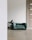 A minimalist room with a green pet bed placed on a light wooden floor next to a potted plant. The door in the background is made of light wood with a modern handle. The overall setting is clean and uncluttered.