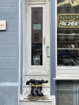 An entrance to a shop with a narrow glass door is visible, accompanied by a window display on the right showing footwear. A Batman costume torso with an emblematic yellow belt is placed on the steps leading to the door. A sign with 'Schuh-Service FACHBERATER' is visible through the window.