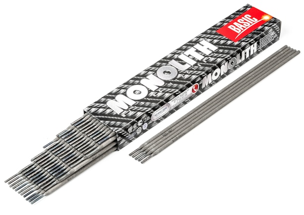 A box labeled 'MONOLITH BASIC' contains a collection of gray welding electrodes. The box has a black and white pattern with a prominent red label. Several electrodes are partially visible, stacked neatly inside the box, while others are laid out side by side in front of it.