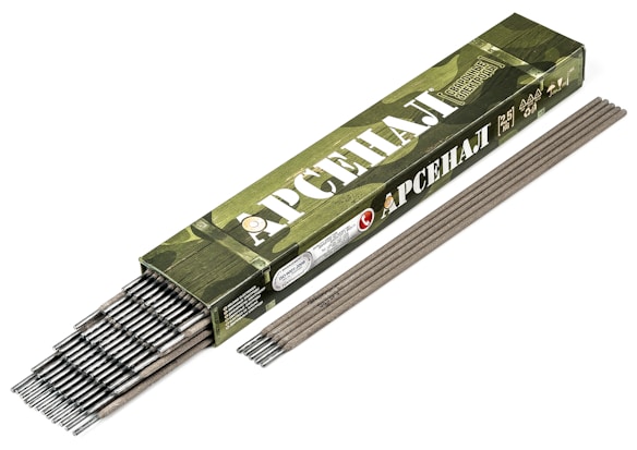 A box of welding electrodes is displayed, with several electrodes partially extending from the open end of the box. The box features a camouflage design in shades of green and is marked with Cyrillic text.