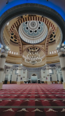 Intricate interior of a mosque featuring a grand dome adorned with geometric patterns and arabesque designs. The ceiling is ornately decorated with chandeliers hanging from above, illuminating the detailed artwork. Arched windows and pillars contribute to the architectural elegance.