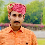 A man wearing a traditional red and white patterned turban adorned with a decorative brooch. He is dressed in an orange shirt and stands against a backdrop of lush greenery and flowers.