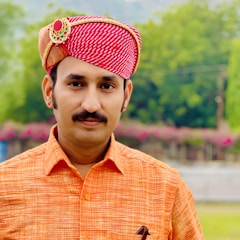 A man wearing a traditional red and white patterned turban adorned with a decorative brooch. He is dressed in an orange shirt and stands against a backdrop of lush greenery and flowers.