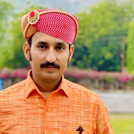 A man wearing a traditional red and white patterned turban adorned with a decorative brooch. He is dressed in an orange shirt and stands against a backdrop of lush greenery and flowers.