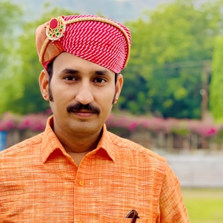 A man wearing a traditional red and white patterned turban adorned with a decorative brooch. He is dressed in an orange shirt and stands against a backdrop of lush greenery and flowers.