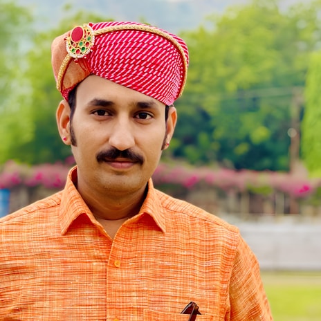 A man wearing a traditional red and white patterned turban adorned with a decorative brooch. He is dressed in an orange shirt and stands against a backdrop of lush greenery and flowers.