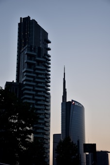 Two modern skyscrapers dominate the skyline against a fading twilight backdrop. The buildings have a sleek and contemporary architectural design, with the structure on the right displaying a logo featuring the name UniCredit. The scene is silhouetted by nearby trees which are partially visible, adding depth to the urban landscape.