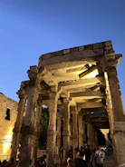 An ancient stone structure with intricately carved columns is illuminated by warm lighting against a backdrop of a deep blue evening sky. People are gathered around the monument, observing and exploring the historical site.