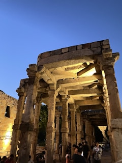 An ancient stone structure with intricately carved columns is illuminated by warm lighting against a backdrop of a deep blue evening sky. People are gathered around the monument, observing and exploring the historical site.