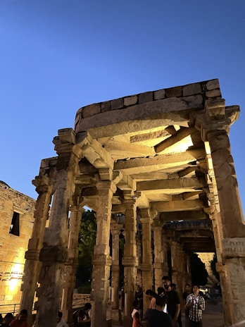 An ancient stone structure with intricately carved columns is illuminated by warm lighting against a backdrop of a deep blue evening sky. People are gathered around the monument, observing and exploring the historical site.