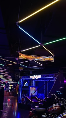 Neon lights in various colors are suspended from the ceiling, creating a geometric pattern. The scene features a vibrant, dimly lit interior with signage that includes 'BROOM WITCH' and arcade-related imagery. There's a motorcycle parked in the foreground and reflections visible on the shiny floor.