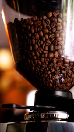 A close-up view of a transparent container filled with dark roasted coffee beans, sitting on top of a coffee grinder. The beans have a rich, glossy appearance, indicating their freshness and quality. The grinder is metallic with a circular adjustment ring at the base, suggesting precise control of grind size.