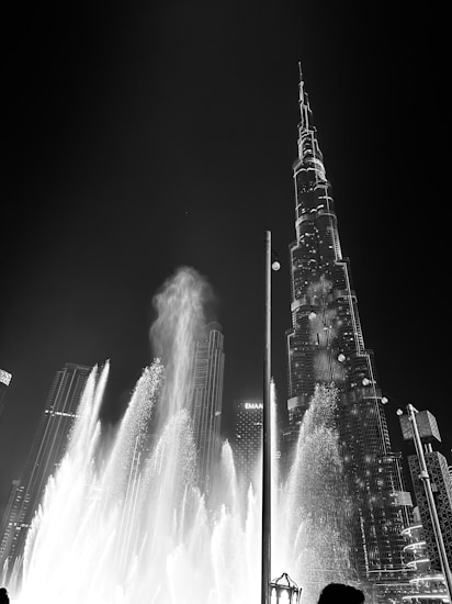 A nighttime cityscape featuring a prominent skyscraper with architectural lighting. Water fountains in the foreground create dynamic, high-reaching sprays. The contrast between the illuminated building and dark sky promotes a sense of grandeur.
