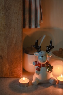 A ceramic reindeer figurine with a red scarf and star ornament sits on a surface surrounded by two lit tealight candles. In the background, there are kitchen items such as a wooden board, a white jar with a lid, and a striped towel hanging. The overall setting suggests a cozy and festive atmosphere.