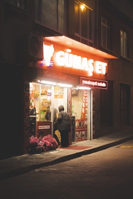 A small illuminated shop is situated on a dimly lit street at night. The shop has a brightly lit sign with the words 'G&uuml;maş Et' glowing in white and orange. A person wearing a hooded jacket stands near the entrance, seemingly looking in or entering the shop. Red and pink baskets or bags are arranged on the sidewalk, adding color to the scene. The street is quiet with no other visible activity.
