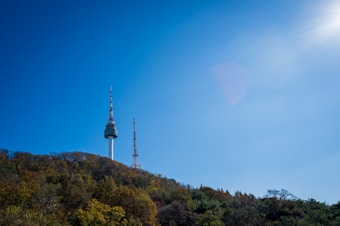 Television and radio towers stand tall on a hill covered with a mix of green and autumn-colored trees under a clear blue sky with the sun shining brightly in the background.