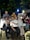 A group of friends pose together while sitting and standing under a tree at night. They are smiling and appear to be having a good time, dressed casually. The setting seems to be an outdoor area possibly at a cafe or restaurant with visible lights and motorcycles in the background.