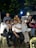 A group of friends pose together while sitting and standing under a tree at night. They are smiling and appear to be having a good time, dressed casually. The setting seems to be an outdoor area possibly at a cafe or restaurant with visible lights and motorcycles in the background.