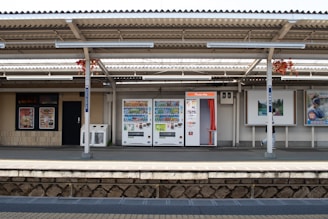 A train station platform featuring vending machines and a photo booth, with advertisement posters on the wall behind. The area is shaded by a corrugated metal roof, with some red foliage visible hanging from the roof beams. The platform is empty except for a person walking by, mostly obscured by a post.
