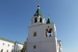 A tall white church with green roofs and bell towers stands against a clear blue sky. It features traditional Orthodox architecture with arched windows, bell openings, and a gold cross perched on top. The building is part of a larger structure with additional white walls and green roofing visible to the left.