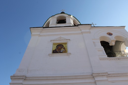 A white church tower under a clear blue sky, featuring an ornate architectural design. A religious icon is visible embedded in the building's facade.