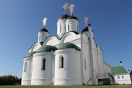 A white orthodox church with multiple green domes topped with golden crosses is surrounded by a clear blue sky. The church features a series of arches and cylindrical towers.