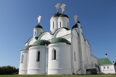 A white orthodox church with multiple green domes topped with golden crosses is surrounded by a clear blue sky. The church features a series of arches and cylindrical towers.