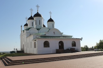 A white Orthodox church building with green roofs and multiple black domes topped with golden crosses. It is situated on a raised platform with brick steps in the foreground. The sky is clear and blue, contributing to a serene atmosphere.