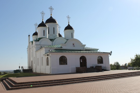 A white Orthodox church building with green roofs and multiple black domes topped with golden crosses. It is situated on a raised platform with brick steps in the foreground. The sky is clear and blue, contributing to a serene atmosphere.