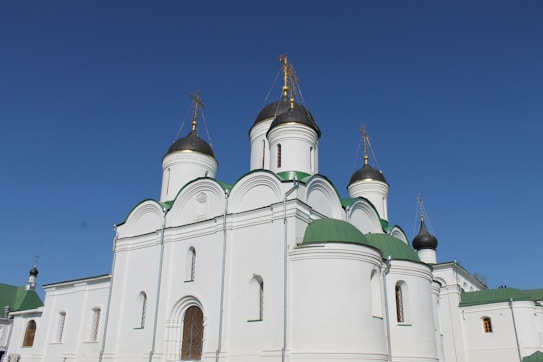 A white orthodox church with multiple dark domes topped with golden cross structures. The building features arched windows and green rooftop elements blending with the pristine blue sky.