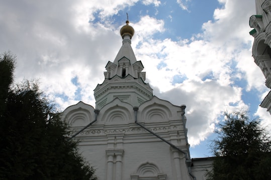 A white, ornate church dome with a gold cross stands tall against a partly cloudy blue sky, surrounded by green trees. The architecture features arches and intricate designs.