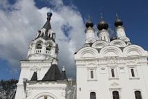 A white orthodox church with ornate architecture, featuring multiple domes in black and gold. The blue sky and fluffy white clouds serve as a backdrop. The structure displays intricate details and patterns characteristic of religious buildings.