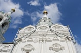 A striking white Orthodox church with intricate architectural details and golden domes set against a bright blue sky. The facade features arched windows and decorative patterns, highlighting traditional Russian design elements.