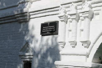 A section of a white brick wall with intricate architectural designs and shadows from nearby trees. There is a dark rectangular plaque with text in Russian, mounted on the wall.