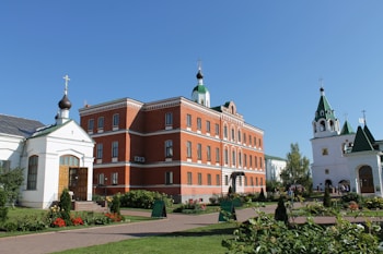 A collection of buildings, including a red-brick building with white accents and green domes topped with crosses, and a white building with green roofs and a tall tower. The buildings are surrounded by a well-maintained garden with flowers, bushes, and paths. The sky is clear and blue.